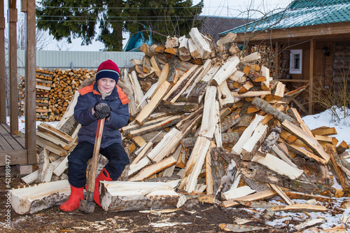 The boy sits next to a pile of firewood and holds a large ax in his hands. Humorous photo.