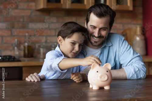Foto Smiling young Caucasian dad and little 7s son manage family budget put coin into piggybank save money for future needs