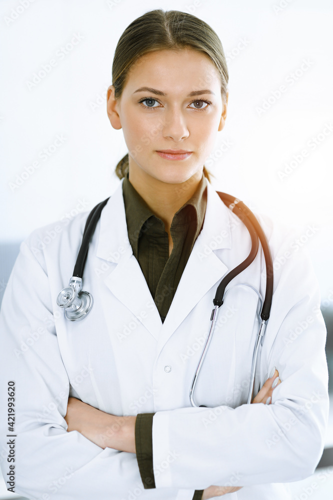 Woman-doctor standing with arms crossed and looking at camera in sunny clinic. Medicine and healthcare Woman-doctor standing with arms crossed and looking at camera in sunny clinic. Medicine and healthcare