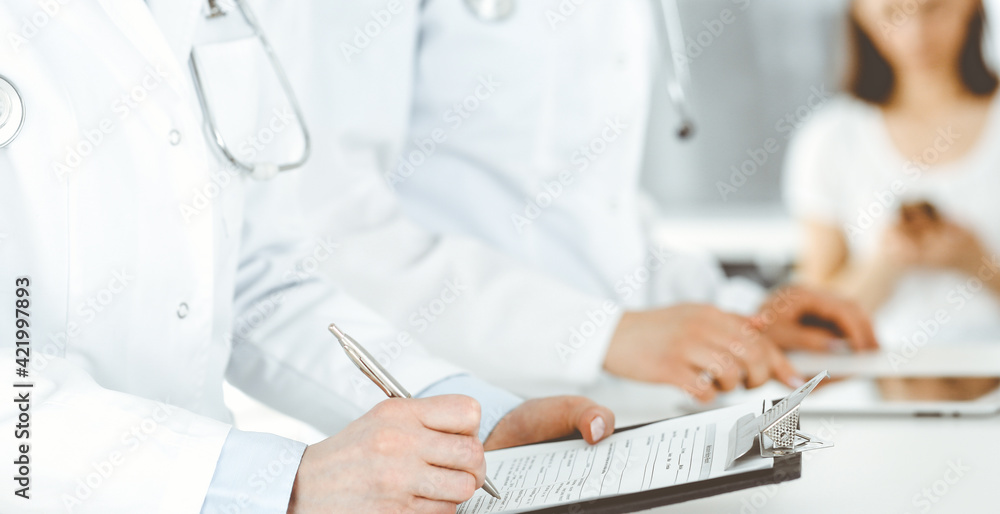 Unknown woman-doctors at work with patient at the background. Female physicians filling up medical documents or prescription while standing in hospital reception desk, close-up. Health care concept Unknown woman-doctors at work with patient at the background. Female physicians filling up medical documents or prescription while standing in hospital reception desk, close-up. Health care concept