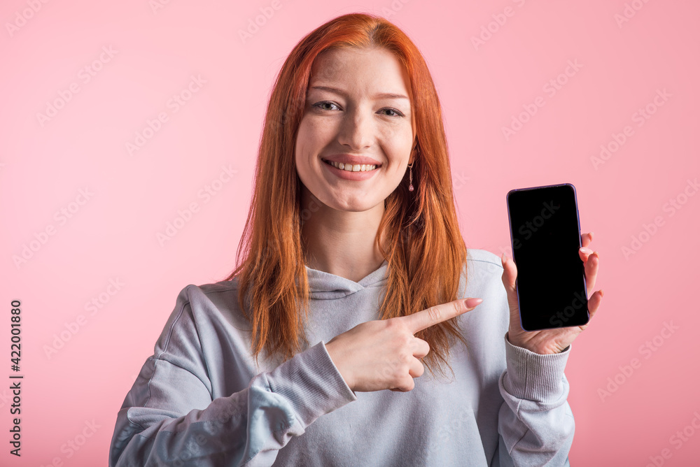Redhead teenager girl points with finger at smartphone blank screen in studio on pink background Redhead teenager girl points with finger at smartphone blank screen in studio on pink background