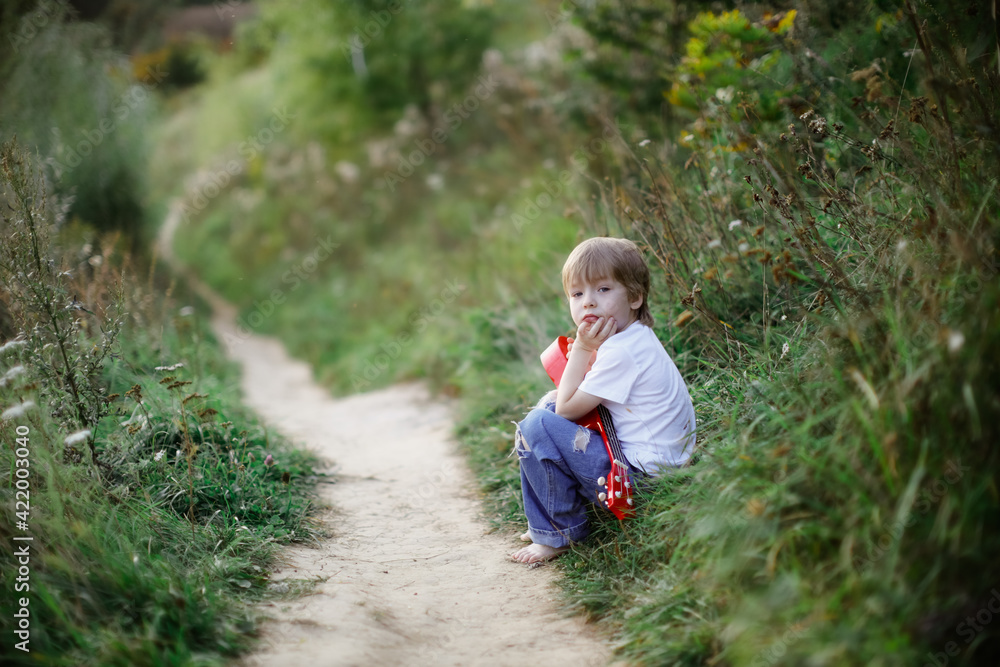 Sad boy child in a white T-shirt sits on the trail with a ukulele, the concept of failure and difficulty, children's emotions. Summer park, Sad boy child in a white T-shirt sits on the trail with a ukulele, the concept of failure and difficulty, children's emotions. Summer park,