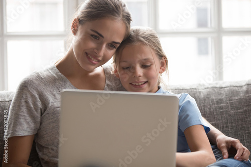 Happy young Caucasian mother and little teen daughter relax at home using modern computer together. Smiling mom and small girl child look at laptop screen talk on video digital webcam call online.