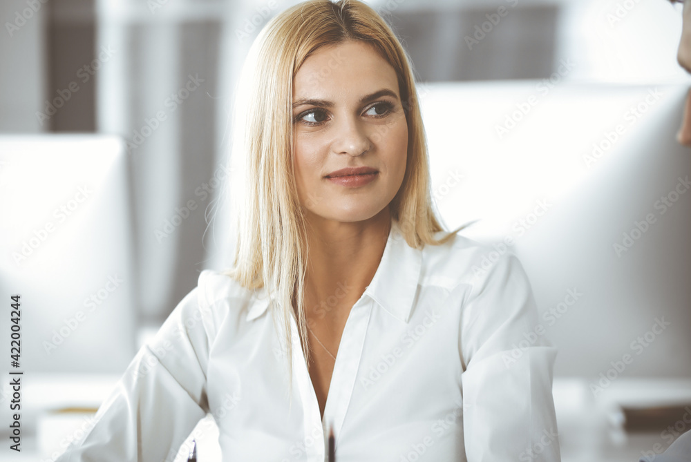 Group of business people discussing questions at meeting. Headshot of blonde businesswoman while smiling to her colleague at office negotiation. Teamwork and cooperation in corporate occupation Group of business people discussing questions at meeting. Headshot of blonde businesswoman while smiling to her colleague at office negotiation. Teamwork and cooperation in corporate occupation