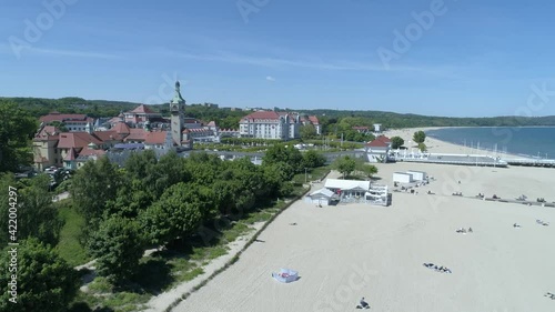 Sandy beach leading straight down to the blue sea. In the middle of the frame there is a boulevard with lots of green trees. In the distance, city buildings and the entrance to the Pier.