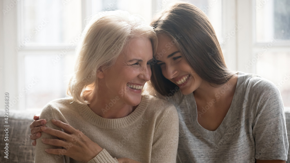 Overjoyed millennial Caucasian woman embrace happy older 60s mom enjoy family weekend reunion together. Smiling mature mother and adult grownup daughter hug laugh sharing secrets at home. - obrazy, fototapety, plakaty 