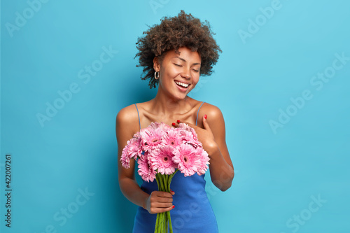 Happy dark skinned pleased woman enjoys romantic date gets bouquet of gerbera flowers from boyfriend closes eyes from pleasure isolated over blue background feels very thankful. Feminity concept