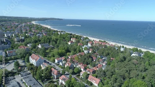 Seaside city landscape from the bird's eye view. In the foreground there are urban buildings with lots of green trees. In the distance, you can see the Baltic Sea and the famous Sopot Pier.