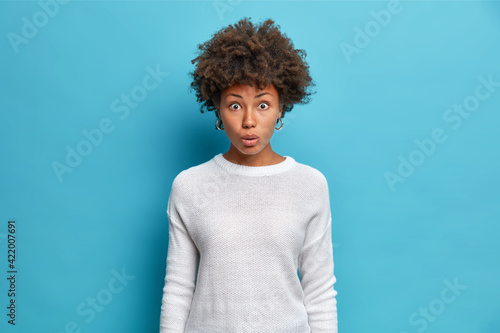Photo of confused shocked beautiful African American woman gazes surprisingly atcamera impresed being accused or blamed wears casual white jumper isolated over blue background. Human reactions
