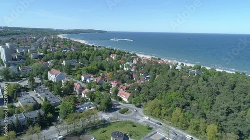 Seaside city landscape from the bird's eye view. In the foreground there are urban buildings with lots of green trees. In the distance, you can see the Baltic Sea and the famous Sopot Pier.