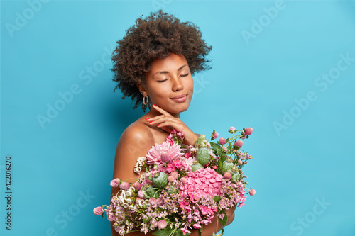 Pleased gentle female model touches jawline closes eyes enjoys lovely moment poses half naked with bunch of beautiful flowers isolated over blue background. Ethnic woman with bouquet poses indoor