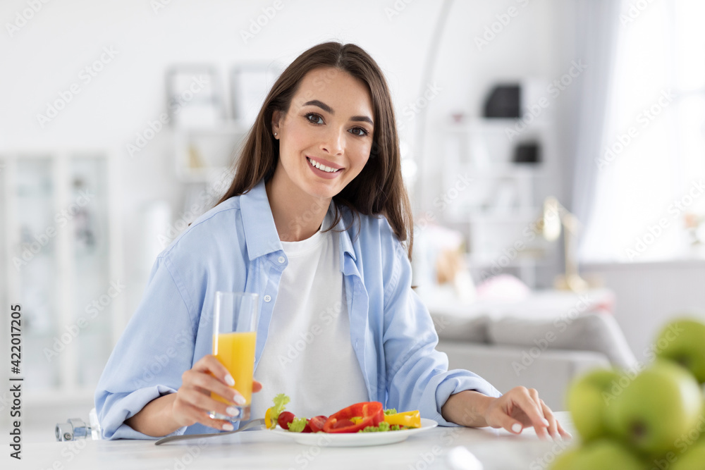 Smiling woman eating wholesome breakfast fresh vegetable salad and orange juice at the kitchen table at home looking at the camera. Smiling woman eating wholesome breakfast fresh vegetable salad and orange juice at the kitchen table at home looking at the camera.