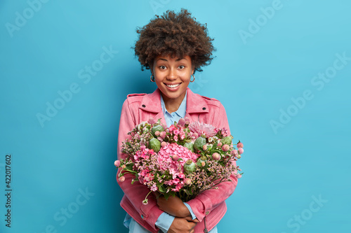 Indoor shot of positive young African American woman embraces big bunch of flowers smiles pleasantly dressed in pink jacket isolated over blue background. Spring portrait. Mothers Day concept