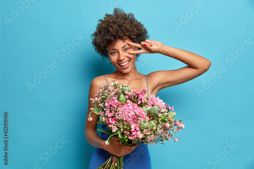 Horizontal shot of cheerful young African American woman shows peace gesture holds big bouquet of flowers smiles broadly has festive mood isolated over blue background. Spring holidays concept