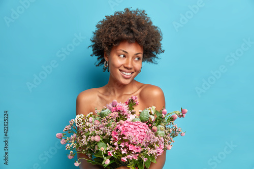 Portrait of natural dark skinned young woman looks aside gladfully smiles gently holds beautiful boquet over naked body poses with bare shoulders isolated over blue backgroun has tender look