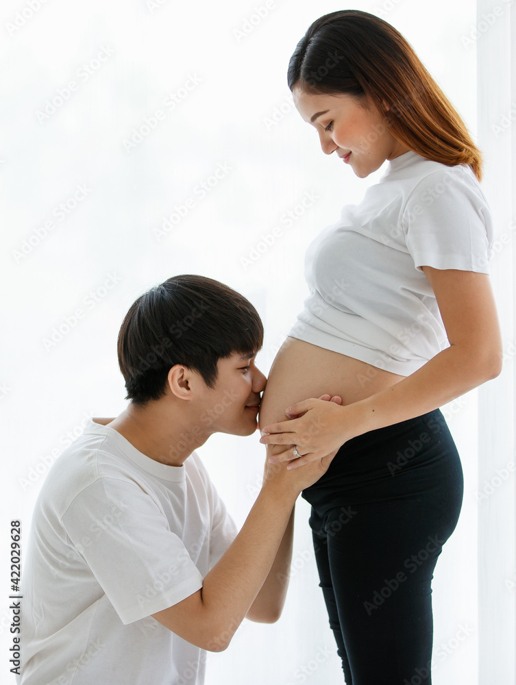 portrait of a very lovely young Asian couple. A husband sitting and kissing a wife's belly while a pregnant woman standing smiling and looking at a man with much love portrait of a very lovely young Asian couple. A husband sitting and kissing a wife's belly while a pregnant woman standing smiling and looking at a man with much love