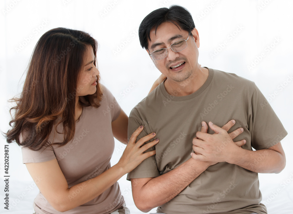 Portrait shot of middle-aged Asian lover couple wearing eyeglasses. Husband abruptly feels uncomfortable from the cardiac chest pain with concerned wife looking and supporting him with care. Portrait shot of middle-aged Asian lover couple wearing eyeglasses. Husband abruptly feels uncomfortable from the cardiac chest pain with concerned wife looking and supporting him with care.