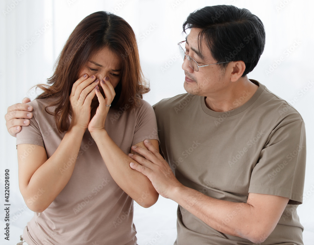 Portrait shot of middle-aged Asian lover couple sitting in the bedroom. Upset wife starts crying because of her health problem with a supportive husband looking carefully and hugging wife with care Portrait shot of middle-aged Asian lover couple sitting in the bedroom. Upset wife starts crying because of her health problem with a supportive husband looking carefully and hugging wife with care
