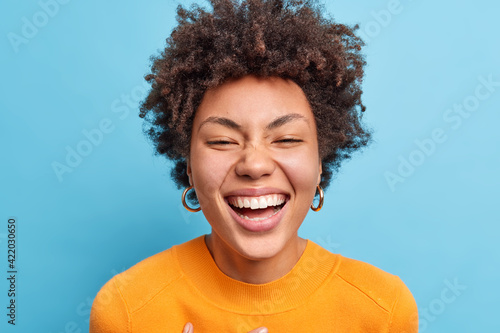 Close up shot of beautiful young African American woman with natural curly hair smiles broadly has positive look at camera being in good mood wears orange jumper isolated over blue background