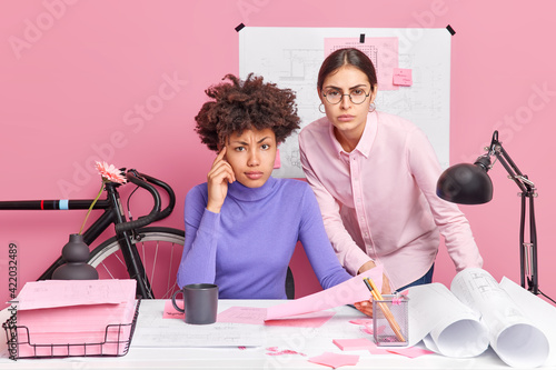 Serious mixed race woman work together on common project in office check documents pose at desktop collaborate and make blueprints isolated over pink background bicyce behind. Teamwork concept photo