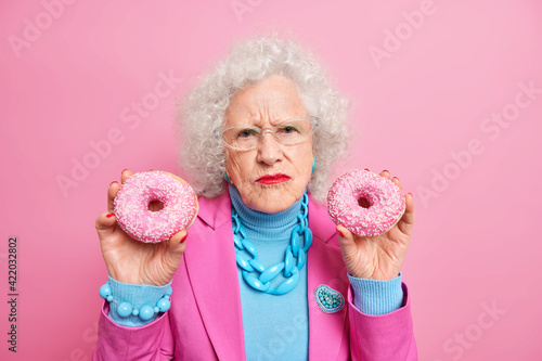 Dissatisfied wrinkled senior woman holds two delicious doughnuts eats junk food wears stylish outfit isolated over pink background. Elderly lady with tasty dessert indoor. Stylish grandmother