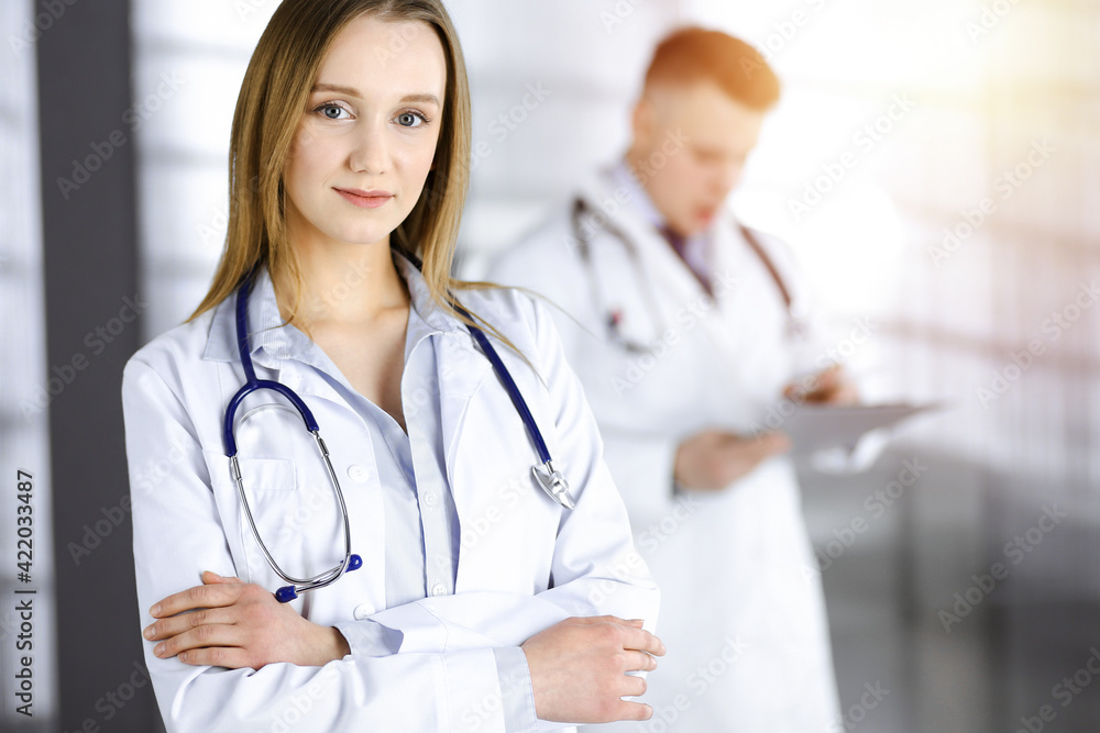Professional beautiful woman-doctor with a stethoscope is standing with crossed arms in a sunny clinic. Young doctors at work in a hospital. Modern medicine and healthcare concept Professional beautiful woman-doctor with a stethoscope is standing with crossed arms in a sunny clinic. Young doctors at work in a hospital. Modern medicine and healthcare concept