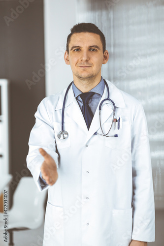 Friendly male doctor standing straight and offering helping hand in clinic near his working place. Perfect medical service in hospital. Medicine and healthcare concept