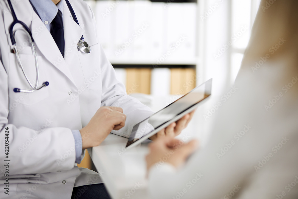 Unknown male doctor and patient woman discussing current health examination while sitting in clinic and using tablet computer. Perfect medical service in hospital. Medicine and healthcare concept Unknown male doctor and patient woman discussing current health examination while sitting in clinic and using tablet computer. Perfect medical service in hospital. Medicine and healthcare concept