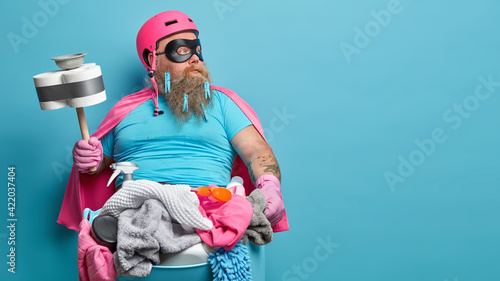 Tableau sur toile Serious bearded man in superhero costume works about house poses with cleaning equipment focused aside holds plunger basin full of dirty laundry isolated over blue background