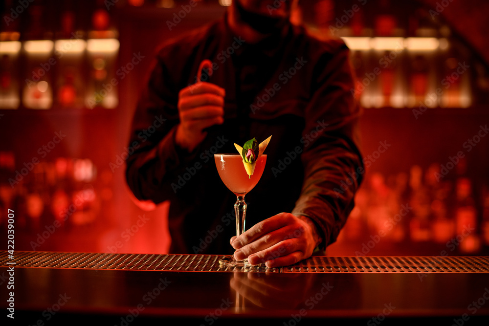 man bartender holds with his hand glass with bright alcoholic cocktail on bar counter.