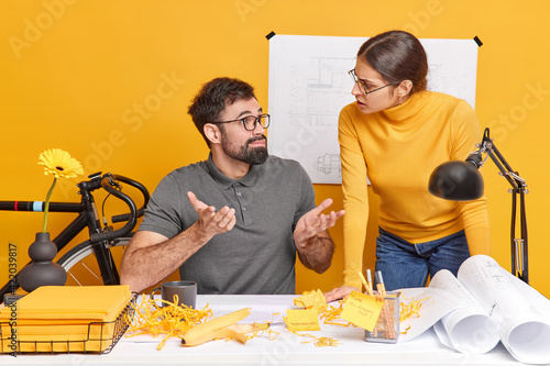 Puzzled man looks at female colleague with confused expression shrugs shoulders doent know how to improve sketch for building project discuss important working issues try to find common solution photo