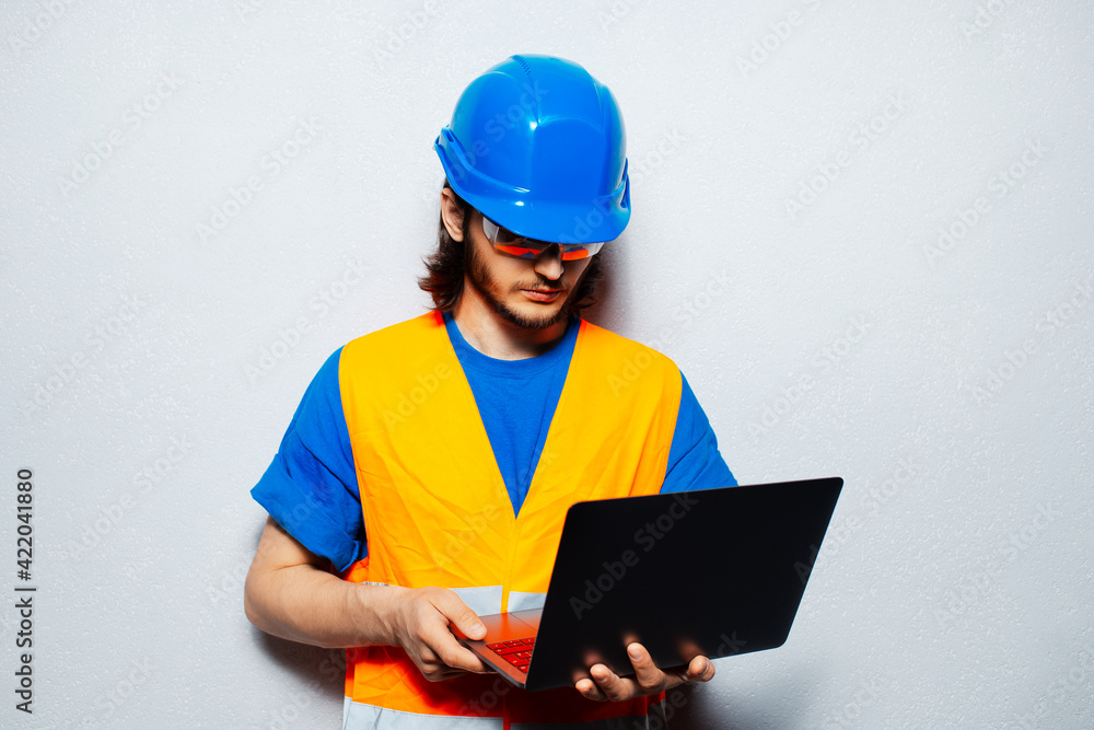 Studio portrait of young construction worker engineer wearing safety equipment, using laptop on background of grey wall. Studio portrait of young construction worker engineer wearing safety equipment, using laptop on background of grey wall.