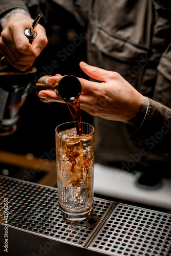 view of glass with ice cubes in which man bartender pours drink from jigger