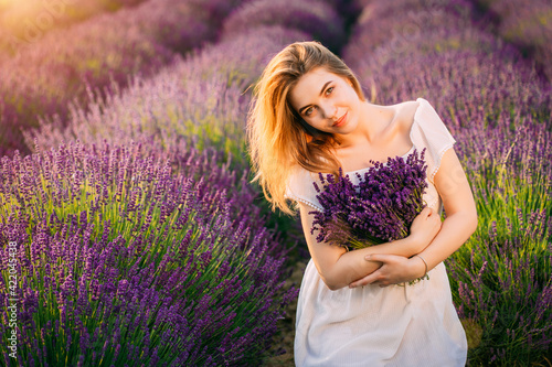 Young and happy blond woman and white dress enjoying spring in a lavender field at sunset holding a bouquet of flowers in her hands