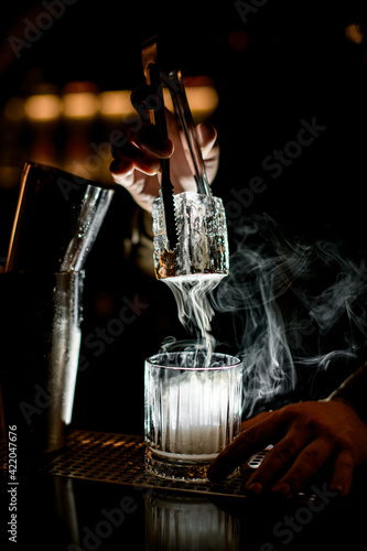 great view of bartender s hand holding large piece of ice using tongs over steaming glass