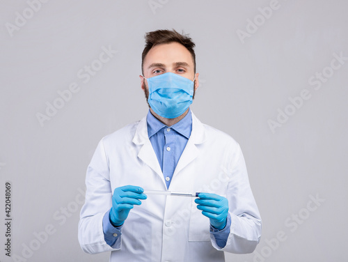 Young doctor wearing medical face mask and rubber gloves  holding coronavirus PCR test tube on grey studio background