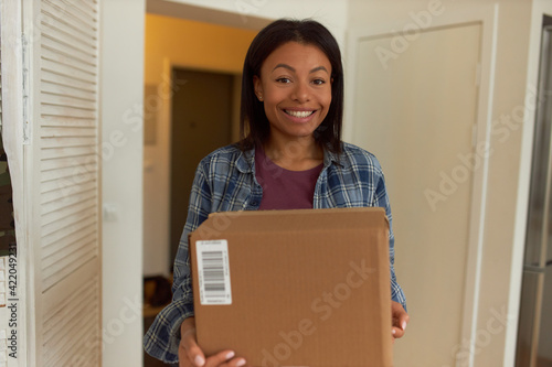 Cheerful dark skinned Hispanic female in casual clothing posing indoors with big cardboard box in her hands packing things moving out to new apartment, having excited joyful facial expression © Anatoliy Karlyuk