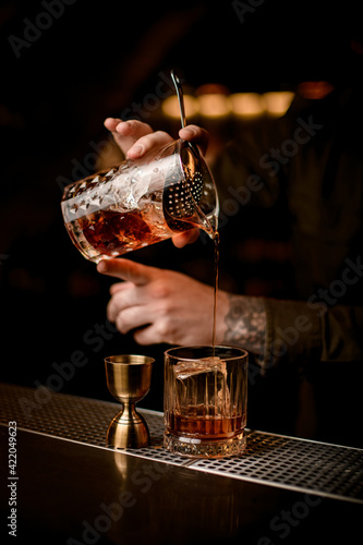 close-up of bartender's hand holds mixing cup and pours cocktail into glass