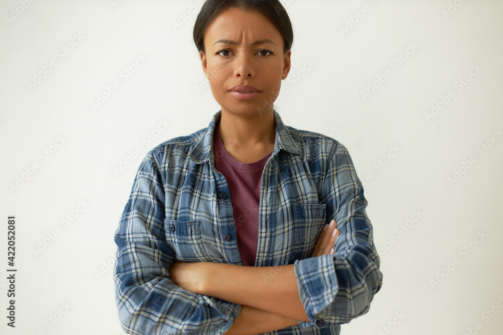 Body language. Isolated shot of grumpy displeased young mixed race woman posing in studio wearing casual plaid shirt frowning her eyebrows, keeping arms folded on her chest expressing disapproval Body language. Isolated shot of grumpy displeased young mixed race woman posing in studio wearing casual plaid shirt frowning her eyebrows, keeping arms folded on her chest expressing disapproval