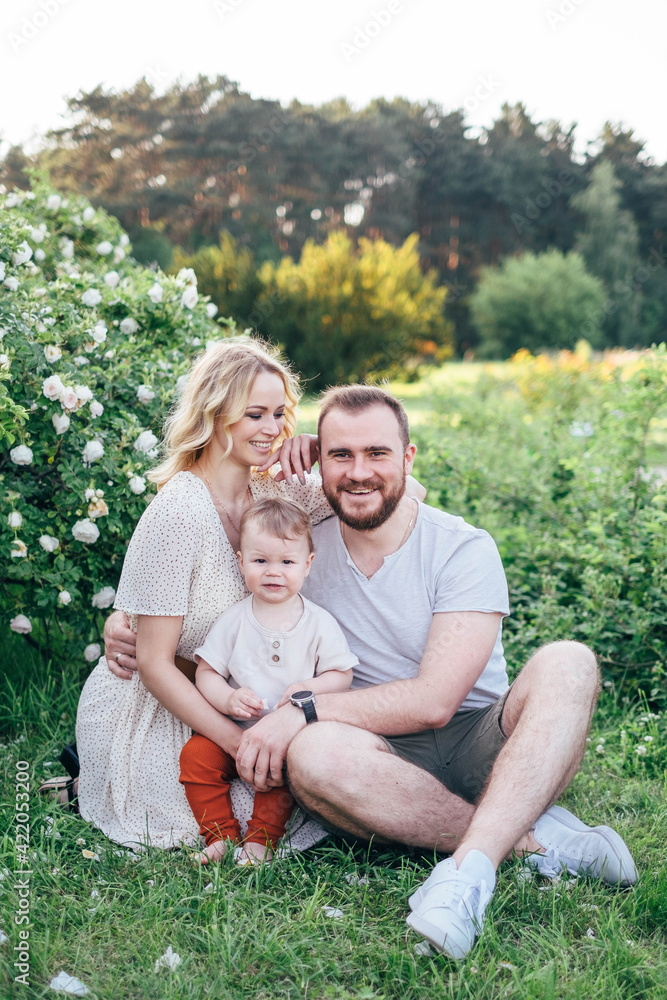Adorable young family with baby boy toddler having weekend in a city park. Blond haired beautiful mom, handsom dad funny kid sitting on grass near blossom roses bush Adorable young family with baby boy toddler having weekend in a city park. Blond haired beautiful mom, handsom dad funny kid sitting on grass near blossom roses bush