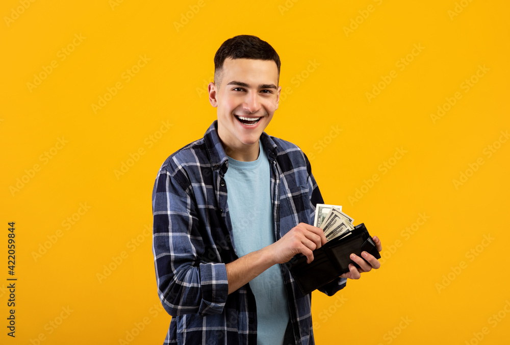 Smiling young man putting money into his wallet on orange studio background Smiling young man putting money into his wallet on orange studio background