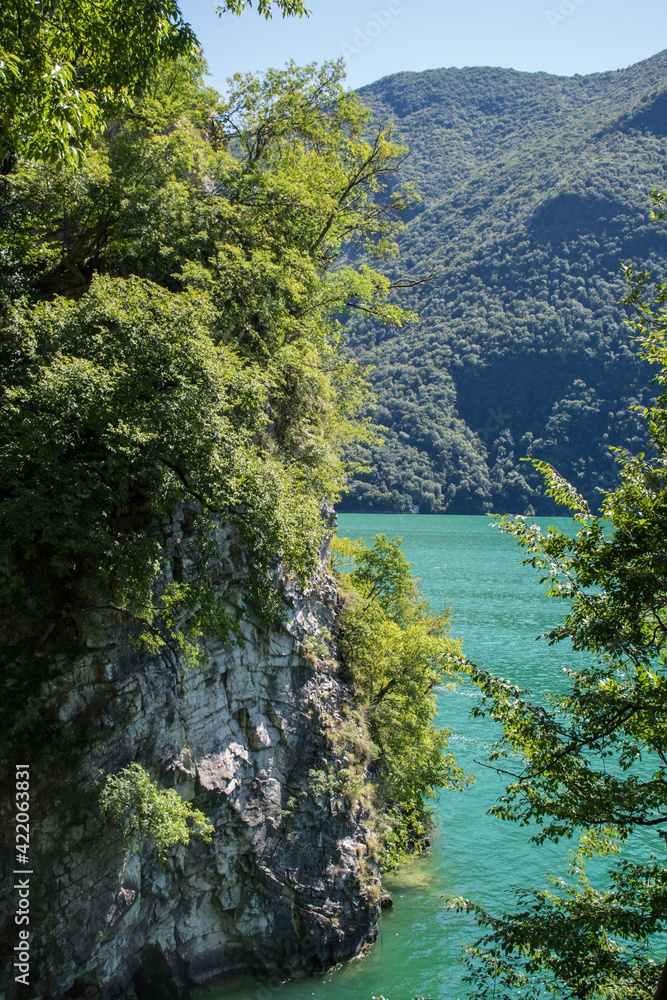 The picturesque rocky shore of the turquoise Lake Lugano in Switzerland is overgrown with forest and shrubs.