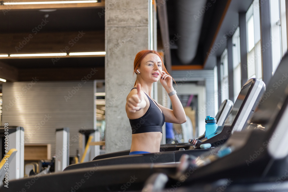 Fototapeta premium Red-haired fitness woman showing thumbs up in modern gym. Happy fit slim woman works out on a treadmill, cardio workout, healthy lifestyle.