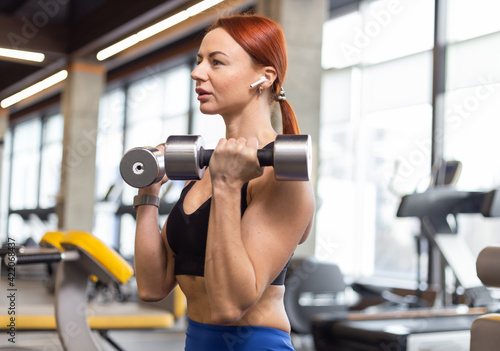 Portrait of a red-haired fitness woman with dumbbells in her hands. Young fit woman working out biceps in modern gym