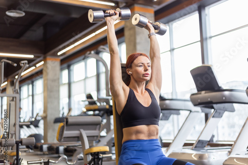 Fit woman trains shoulders with dumbbells in her hands doing dumbbell bench press over her head while sitting on a bench in the gym