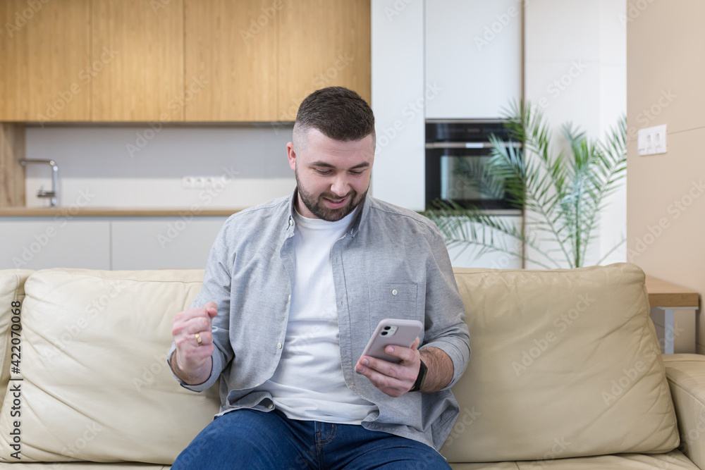 Smiling young man holding mobile phone while sitting on a sofa at home and celebrating Smiling young man holding mobile phone while sitting on a sofa at home and celebrating