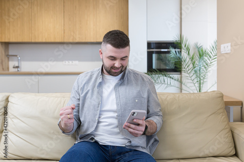 Smiling young man holding mobile phone while sitting on a sofa at home and celebrating