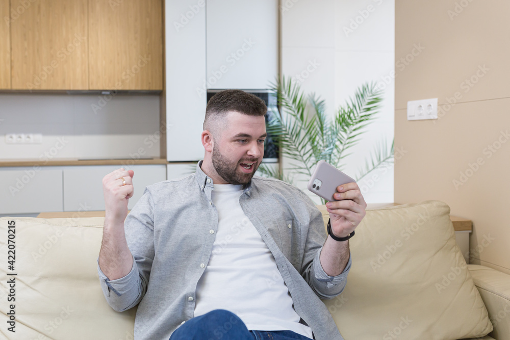 Fototapeta premium Smiling young man holding mobile phone while sitting on a sofa at home and celebrating