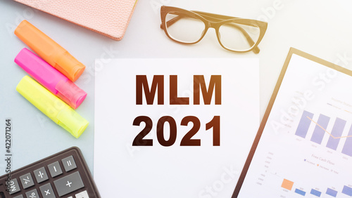 The text MLM 2021 - multilevel marketing on office desk with calculator, markers, glasses and financial charts.