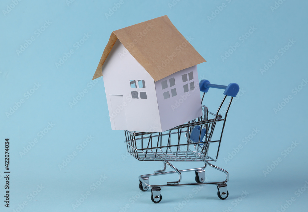 Home purchase. Mini paper house figurine in supermarket trolley on blue background. Home purchase. Mini paper house figurine in supermarket trolley on blue background.